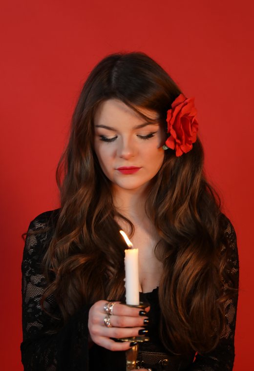 Woman with her eyes lowered holding a lit candle, a red rose in her hair